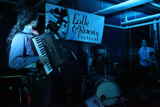 A band playing in a dark bar with a "Folk & Roots Festival" banner hanging in the back.