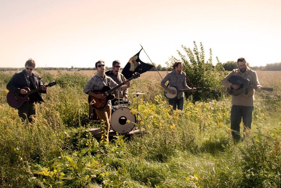 a photo of a band playing in a field. The band consists of five members, each playing a different instrument, including a guitar, a bass guitar, a drum set, a banjo, and a flag. The band members are standing in a line, facing the camera. The field is full of tall grass and wildflowers, and the sky is clear and blue. The photo has a warm, summery feel to it.