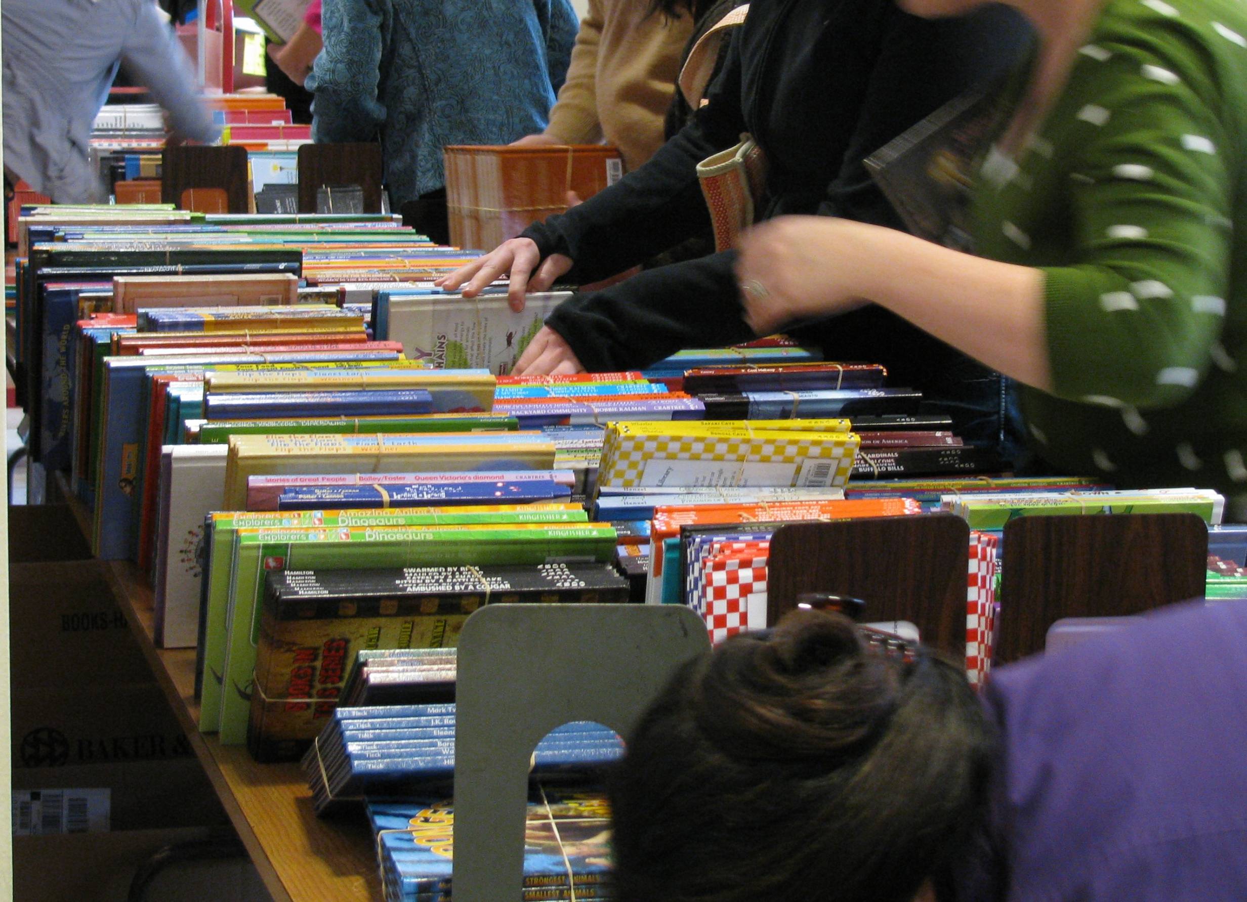 rows of books sit on a table a white women in a green shirt goes through the titles