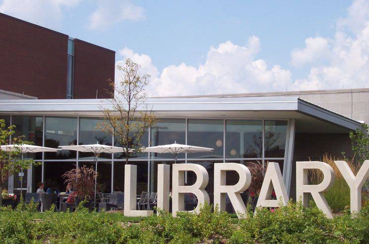 An outside shot of Champaign Public Library. There is a blue sky with clouds; the large brown building with many windows. Huge letters in the grass spell out Library