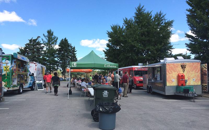 A parking lot with a tent in the middle, and food trucks on the edges and people walking through the parking lot.