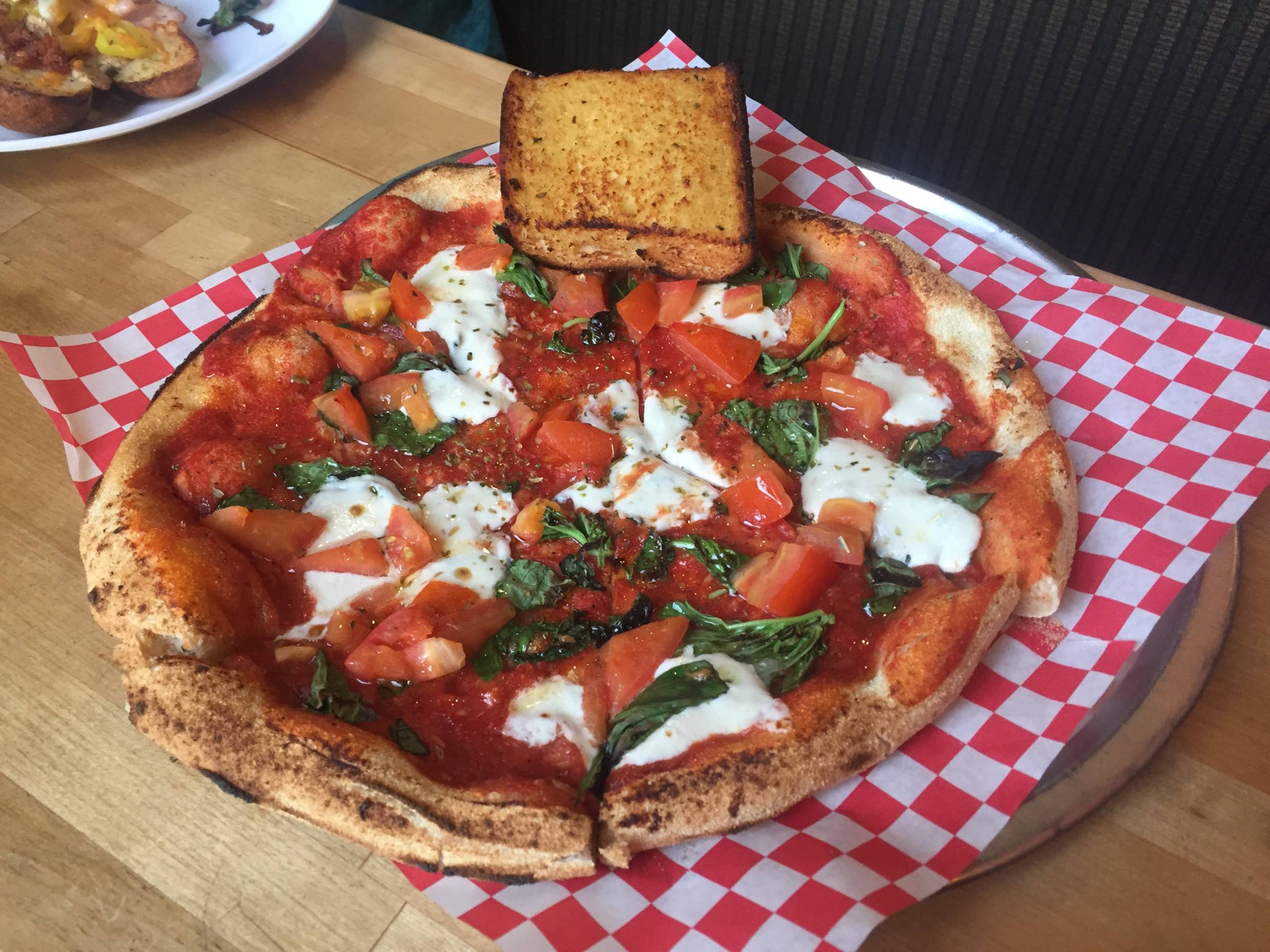 An individual-sized margherita pizza with a slice of garlic bread balanced on the crust. It is served on red and white check paper.