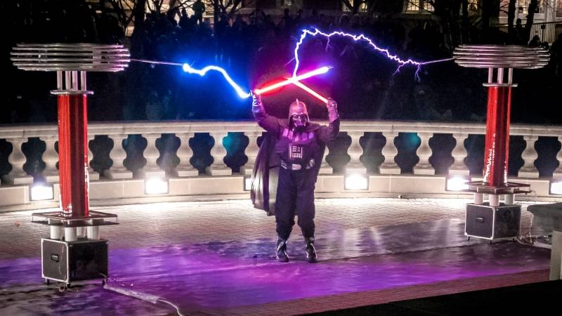 A person dressed as Darth Vader is holding two light sabers, as electric current streams from each.