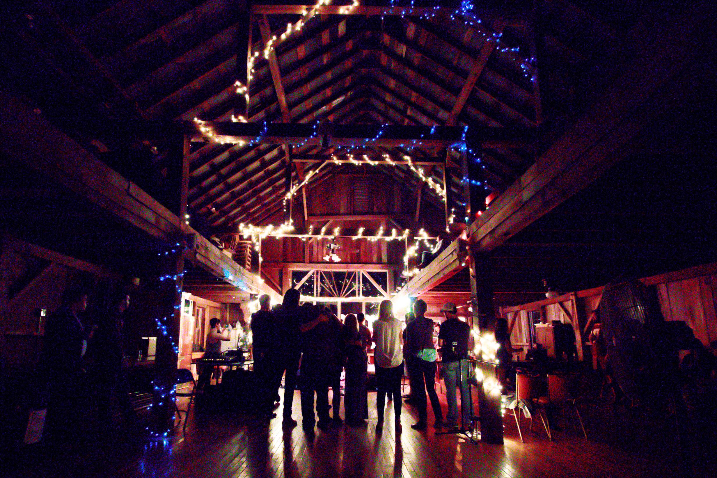 The inside of a barn venue at night during a concert. Lights come from the stage in the background of the image. A crowd is silhouetted in front of the stage.