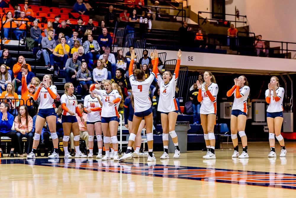A group of volleyball players wearing a uniform of navy shorts, white knee pads and shoes, and white jerseys with orange sleeves and numbers clap and cheer on the court. The stands are full of fans watching.