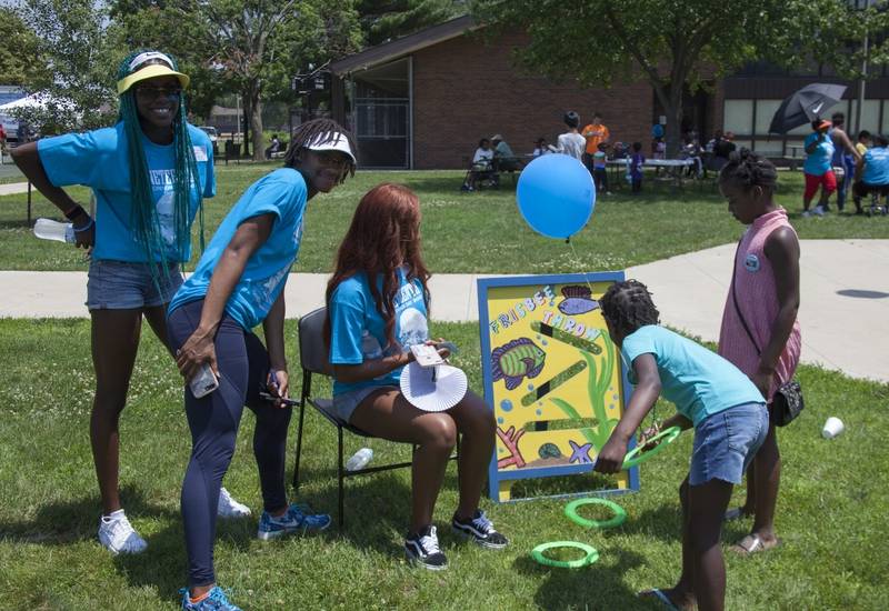 Three young Black women in bright blue t-shirts are watching a young Black boy tossing a bean bag into a board with holes cut out of it.