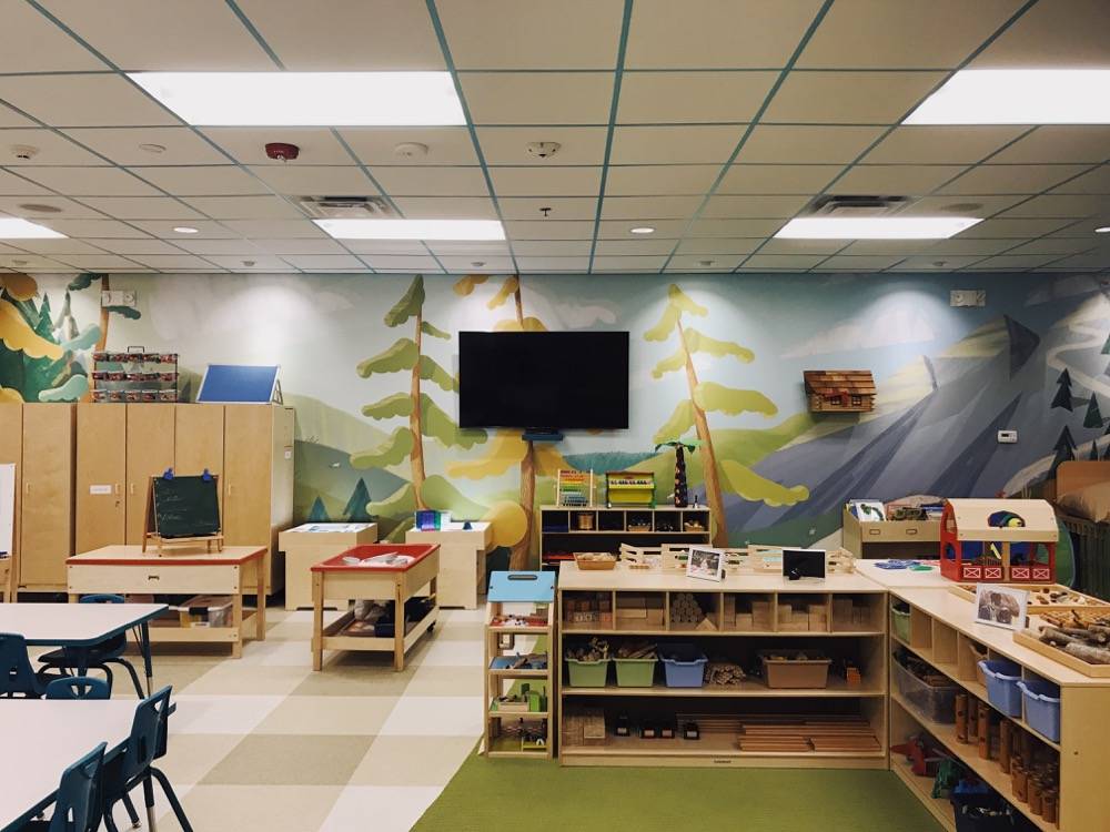 A view of Lodgic's child care center. A mural of a mountain and trees is on the back wall. There are cubbies and tables and kids toys on shelves. A television is on the wall with the mural.