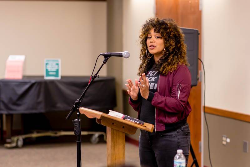 Elizabeth Acevedo stands behind a podium with a microphone. She has curly brown hair and is wearing a black t shirt and maroon jacket