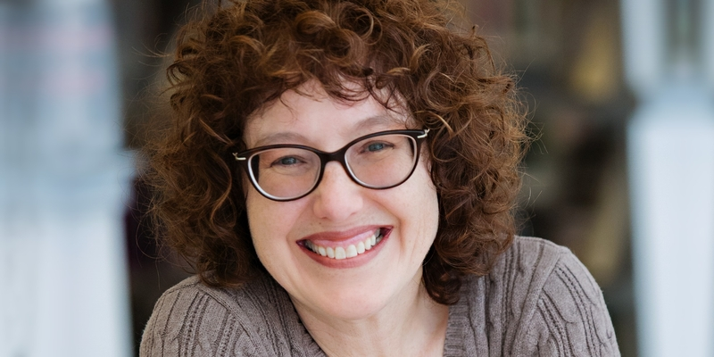 Headshot of a white woman with short curly brownish red hair. She is wearing glasses and a beige sweater.