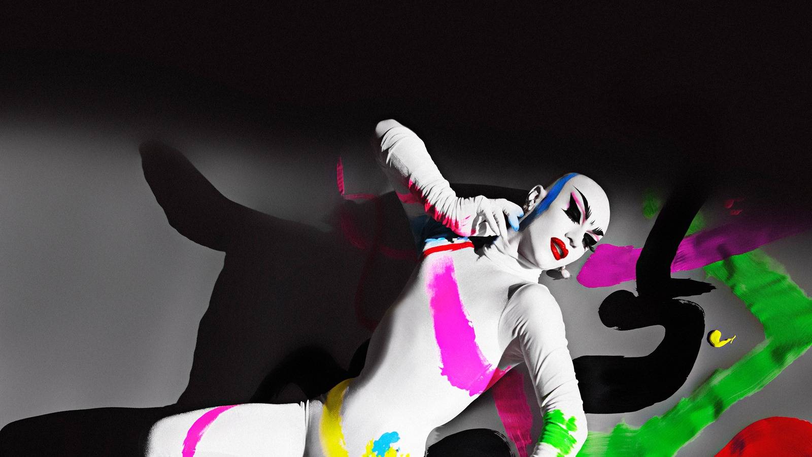 Sasha Velour lays on stage, propped on one arm. She is in an all white bodysuit, and her face is also painted white. Large streaks of neon paint are across the floor and her bodysuit. 