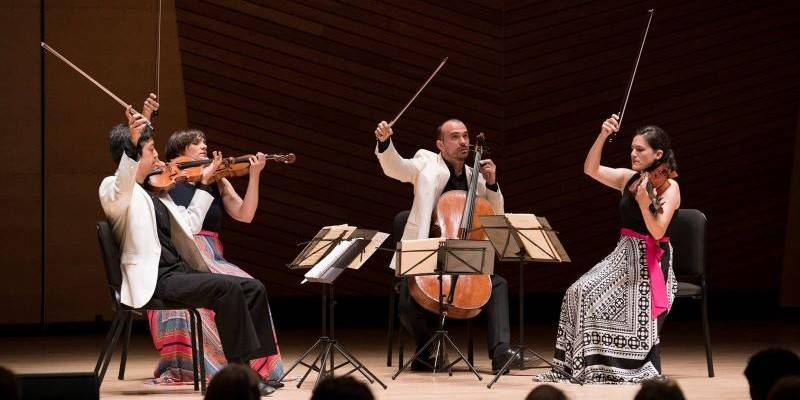 Jupiter String Quartet performing on stage. On the left a man and a woman play violin. In the middle a man plays cello. On the right a woman plays violin. The men are wearing white jackets.