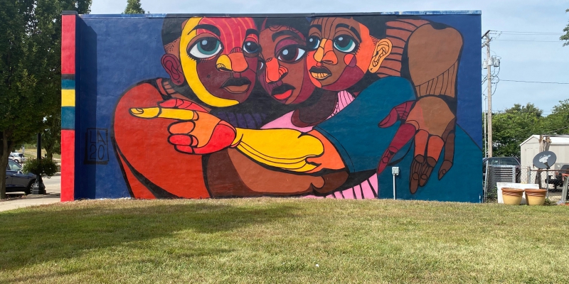 A brightly colored mural covering the side of a building, featuring three African American people embracing, as one points to something unseen.