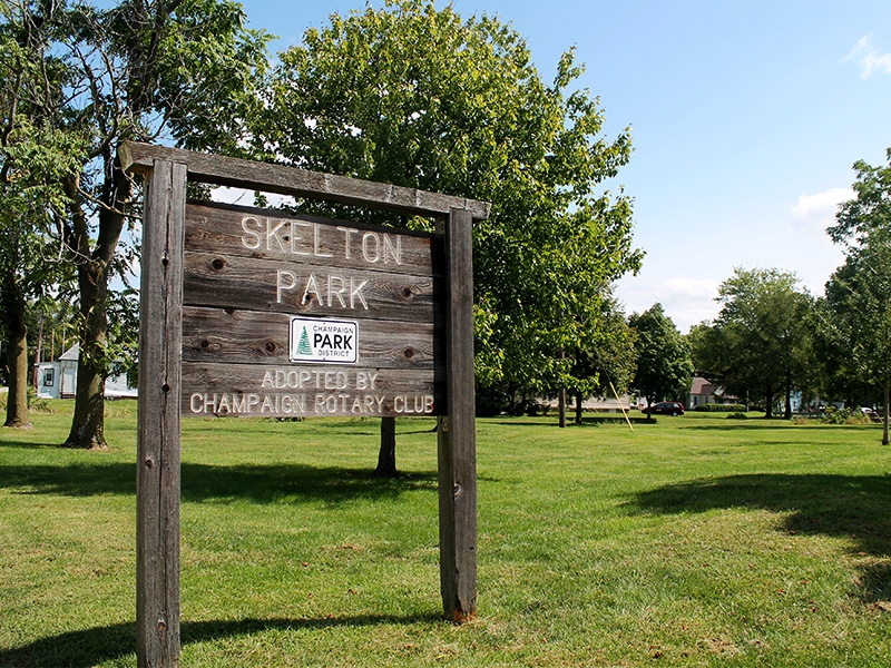 A wooden sign has the words Skelton Park carved into it. It's standing in a field of grass, with leafy green trees in the background.