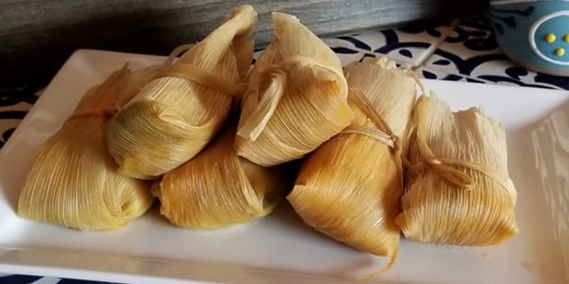 A rectangular white plate has five wrapped tamales stacked on it.