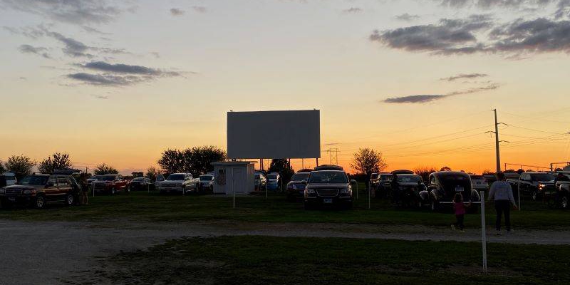 The movie screen at Harvest Moon Drive In, Gibson City IL seen as dusk. The isn't anything on the large, white screen. There are many cars in the lot, and the sky is an orange sunset.