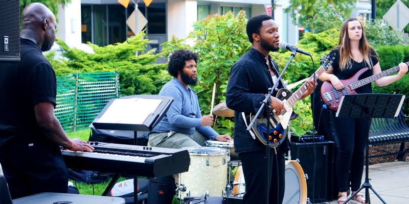 This photo is a photo-realistic image of a band playing in an outdoor setting. The band consists of four members, all playing different instruments including a keyboard, drums, guitar and bass guitar. The band is set up on a concrete patio with a green lawn and trees in the background. There are black and yellow triangular flags hanging above the band. The band is playing in front of a building with large windows.