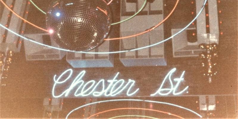 An old photo of the interior of C-Street. There is a blue neon sign that says Chester Street in script. Strings of lights and a disco ball hang above it.