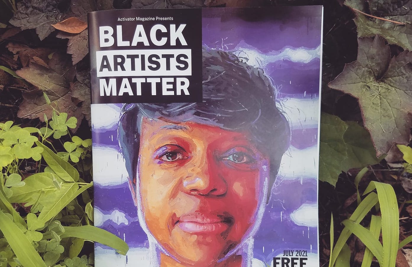 The July 2021 issue of Activator Magazine is a Black Artists Matter issue. The cover features an in-color print of a portrait of Endalyn Taylor by Patrick Earl Hammie. It is a Black woman viewed straight on. She has short, dark hair and looks out at the viewer. The background is purple and white, and there is purple reflected on her face. The magazine is propped up in some grass and leaves.