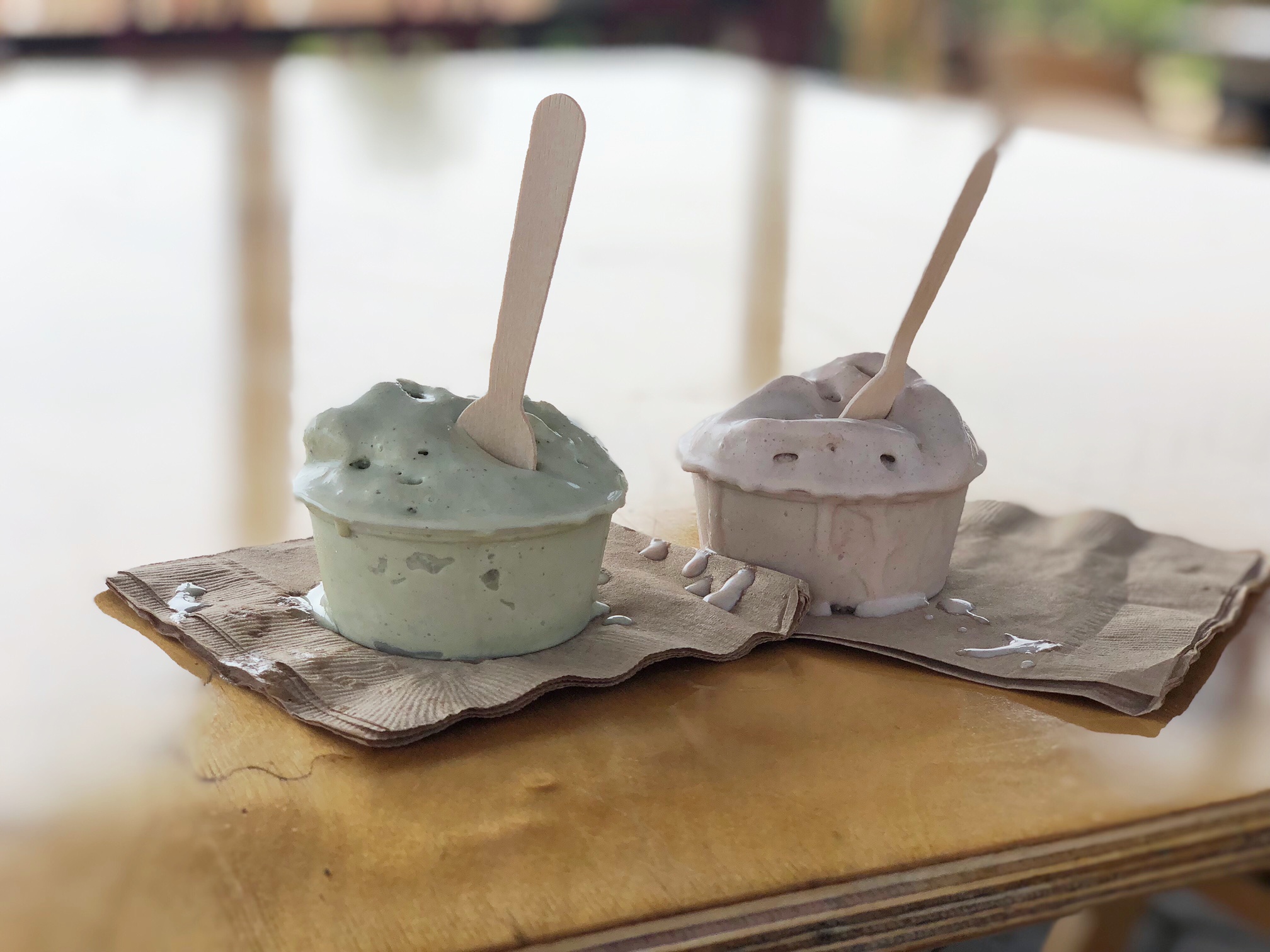 Two small cups of ice cream sitting on napkins on a wooden table. They each have a spoon sticking out of them.