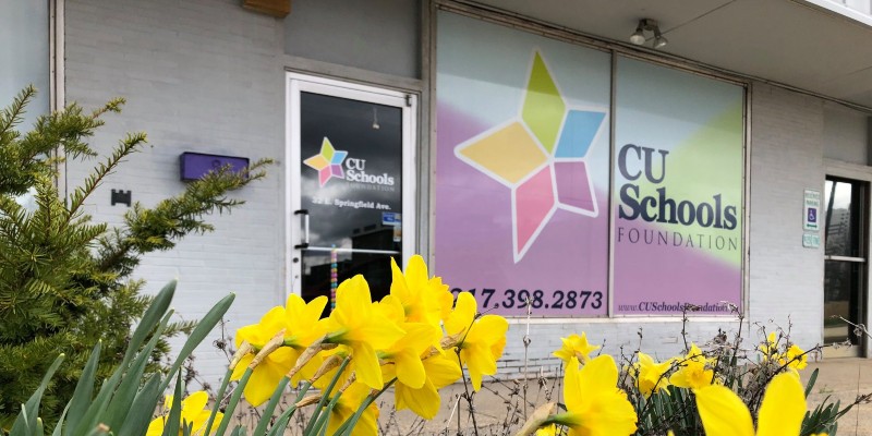 The outside of a building. There is a glass door, then windows covered in a multi-colored sign that says CU Schools Foundation. There are yellow daffodils in the foreground.