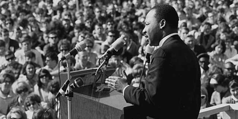 Black and white photo of Dr. Martin Luther King Jr. standing at a lecturn, speaking to a large crowd.
