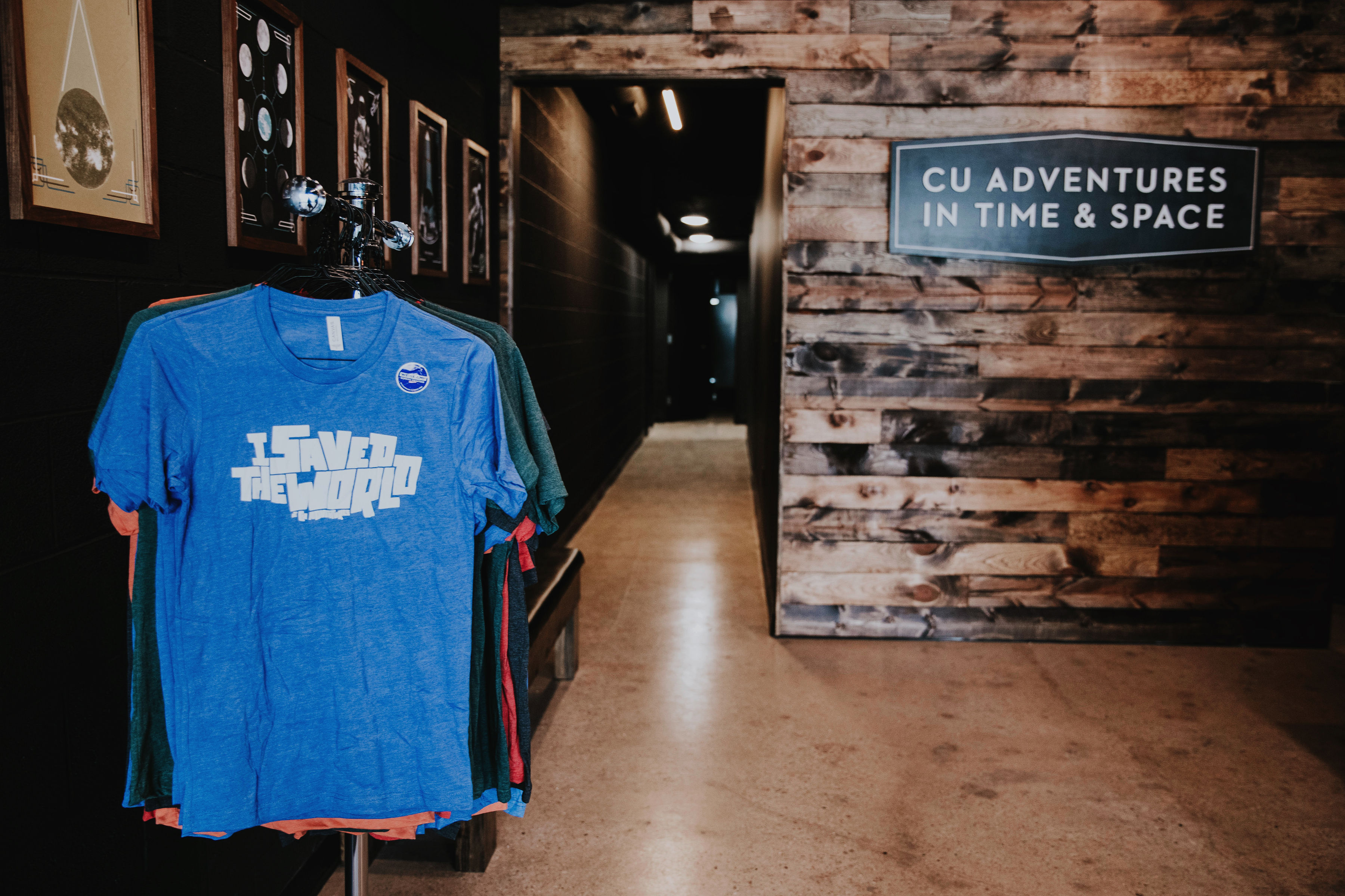 A rack of t-shirts has a blue t-shirt hanging at the front. To the right is a wall with wood slats and and black sign that says C-U Adventures in Time & Space