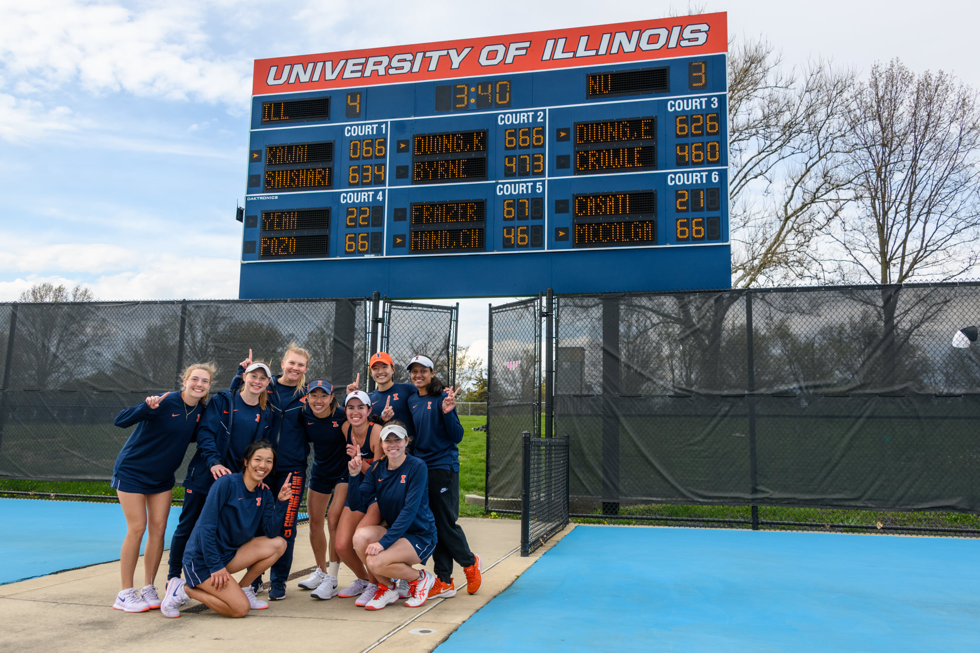 Group of nine women huddled together and smile at the camera. They are all holding up one finger to indicate #1 status. They are standing on a tennis court, with a scoreboard indicating their win behind them. Photo from the Fighting Illini Womenâ€™s Tennis Facebook page.