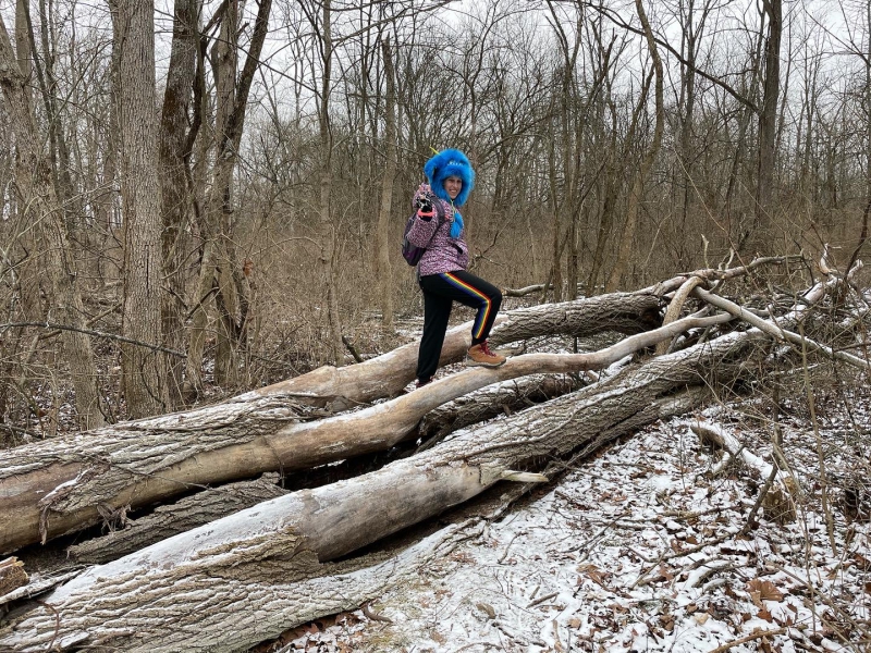 The writer is standing on a tree trunk that is laying on the ground. She's wearing a big blue fuzzy hat, pink jacket, and black pants with rainbow stripes. There is a row of trees in the background. Photo by Mara Thacker.