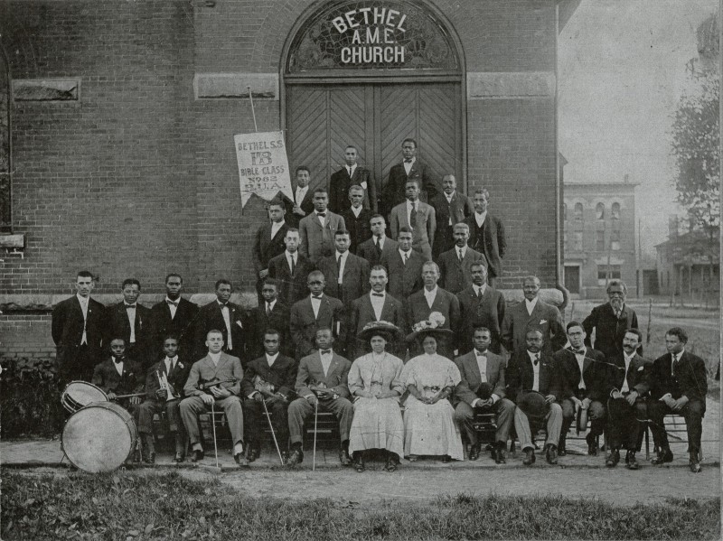 A black and white photo of a group of mostly Black men in suits and two Black women in dresses and hats. They are posed in front of a church, a brick building with a sign over the door that says Bethel AME Church. The man on the far left is sitting behind a drum set, and one person is holding a banner sign that says Bethel S.S. Bible Class. Photo from Facebook event page, courtesy of Doris K. Wylie Hoskins Collection, at the Museum of the Grand Prairie.
