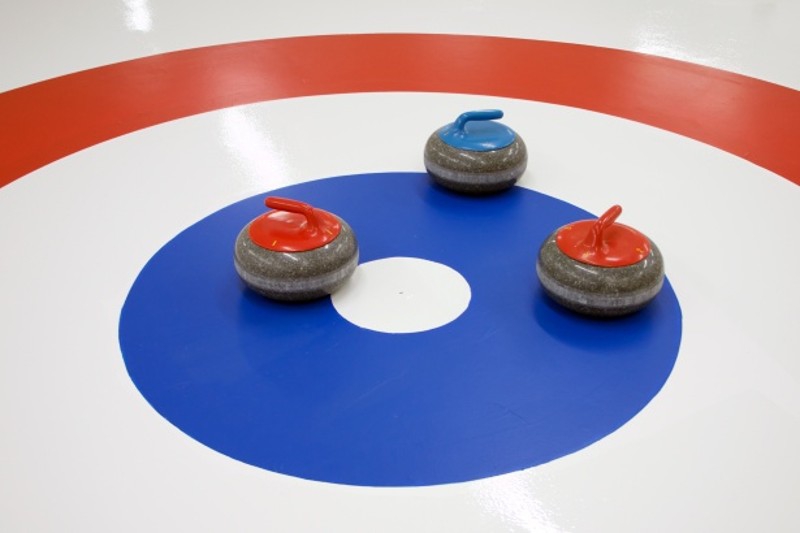 Three curling stones sit in the center of a target on the ice. There is a portion of a red circular band, a white space, then a thick blue circular band with a small white circle in the center. Photo from Prairielands Curling Facebook page. 