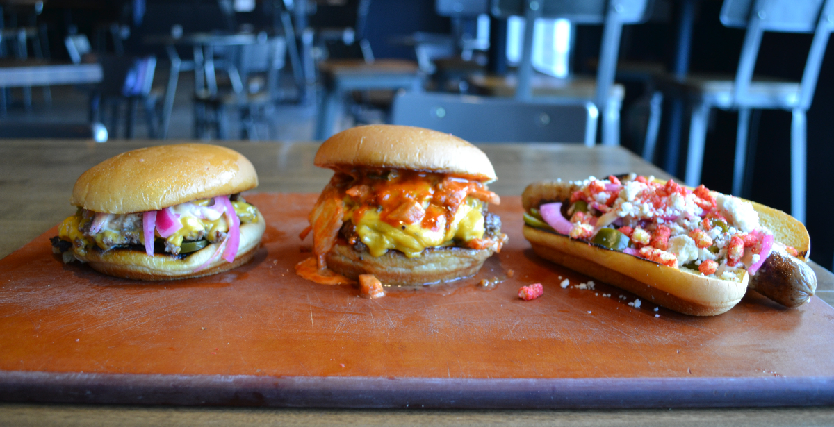 Two burgers and a hot dog sit on a wooden cutting board on a silver table in an empty Champaign bar. The leftmost burger has melted orange cheese and purple pickled onions. The burger in the middle is overflowing with kimchi, and the hot dog is topped with cojita cheese, flaming hot Cheetos, and pickled jalapenos. Photo by Alyssa Buckley.