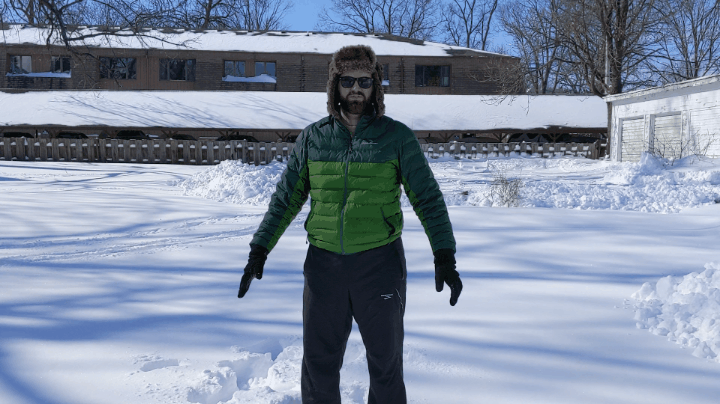 A man in a green puffy jacket, a furry hat with ear flaps, sunglasses, black gloves, and black snow pants is standing in the snow, staring at the camera. Photo by Andrea Black.