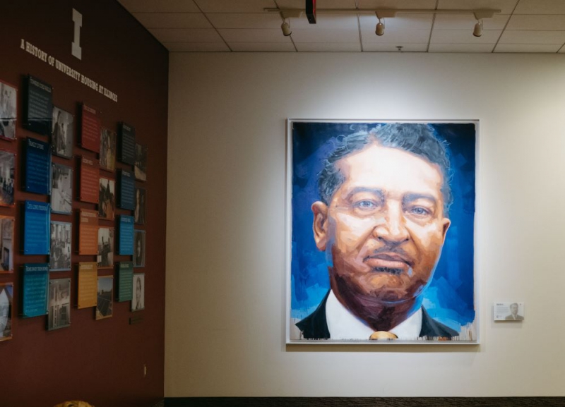 A large portrait of an African American man wearing a white collared shirt and suit jacket. It has a blue background, and hangs on a wall adjacent to a wall with panels about the history of U of I housing. Photo by Anna Longworth.