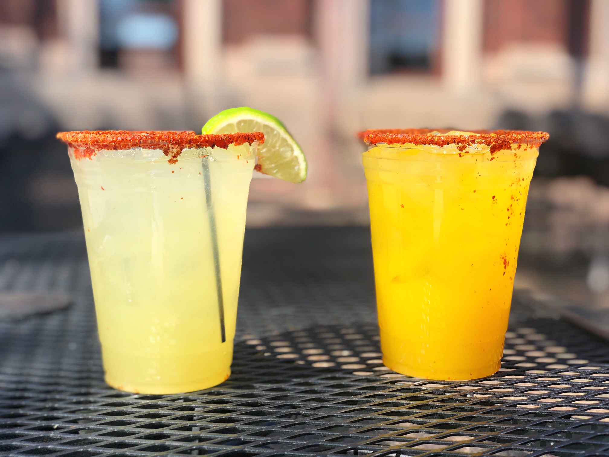 Two margaritas with tajin on the rim, sitting on a black patio table. One is orange in color and one is yellow.