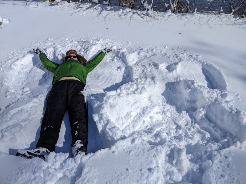The author is laying down in the snow, arms and legs outstretched, making a snow angel. There is a spot next to him where someone has already made a snow angel. Photo by Andrea Black.