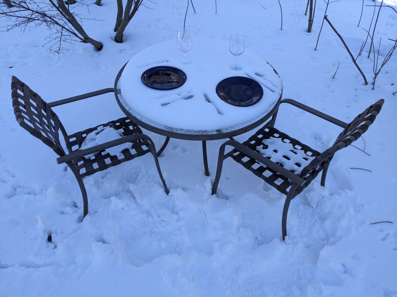 A snow covered round table and two chairs sit in the snow. There are two plates and two wine glasses on the table. Photo by Tom Ackerman.