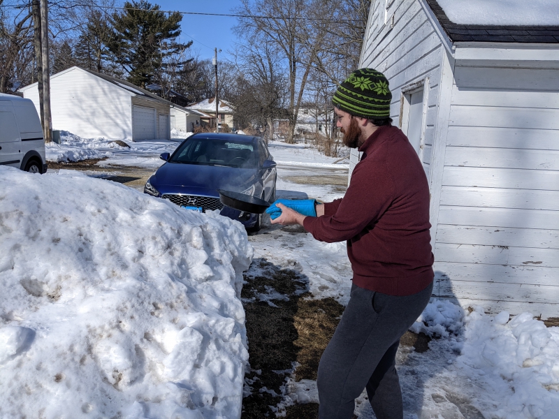 The author is outside wearing a stocking cap, red sweater, and black pants. He is standing next to a large pile of snow, holding a black pan. Photo by Andrea Black.
