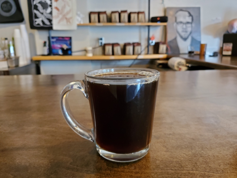 A glass mug filled with black coffee is sitting on a wooden counter top. There are shelves with bags of coffee beans in the background, as well as several art prints. Photo by Rafay Khan.