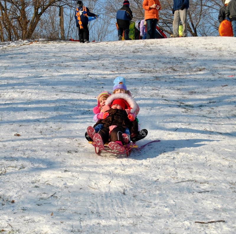 Three bundled up children are on a sled, sliding down a snowy hill. At the top of the hill there are kids and adults in winter gear, some holding sleds. Photo from Champaign Park District website.