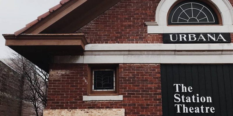 A close up of one corner of The Station Theatre building. It's a brick building with a pointed roof. There is a small square window and a semi-circle window. There is a sign under the latter that is black and says Urbana in white letters. Below that there is a black sign that says The Station Theatre in white letters. Photo by Patrick Singer.