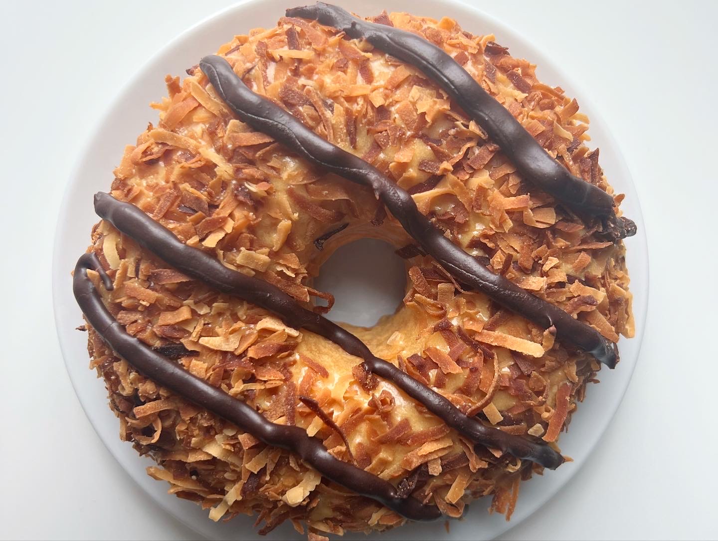 On a white plate on a white table, there is a Girl Scout Samoa cookie-inspired yeast donut with toasted coconut and chocolate drizzle, just like the cookie. Photo by Alyssa Buckley.