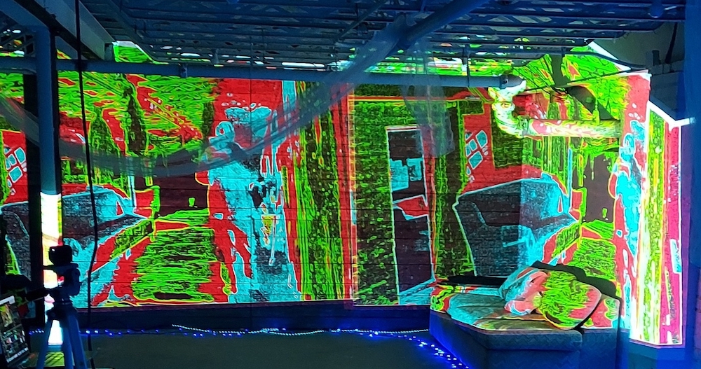 A brick wall with a brightly colored image projected onto it. It appears to show a room with a couch and a door, in red, chartreuse, and light blue. Image from Soundscape's Facebook page. 