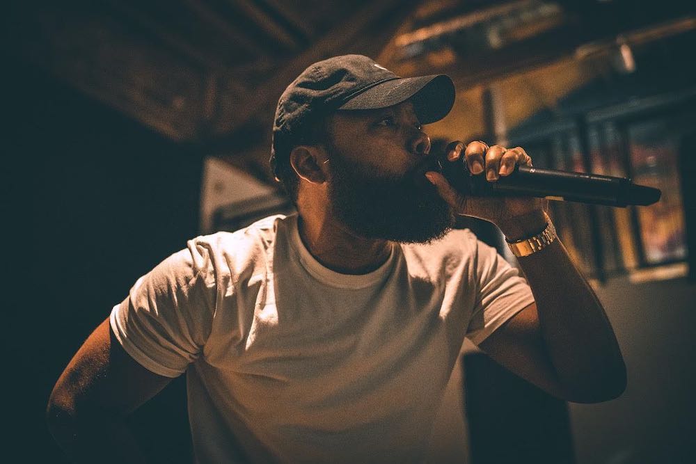 Action shot of rapper Landon Wordswell rapping into a microphone. He is Black man with black baseball cap, full beard, and white t-shirt. Photo by Keshia Murdoch from Landon Wordswell's Facebook page.