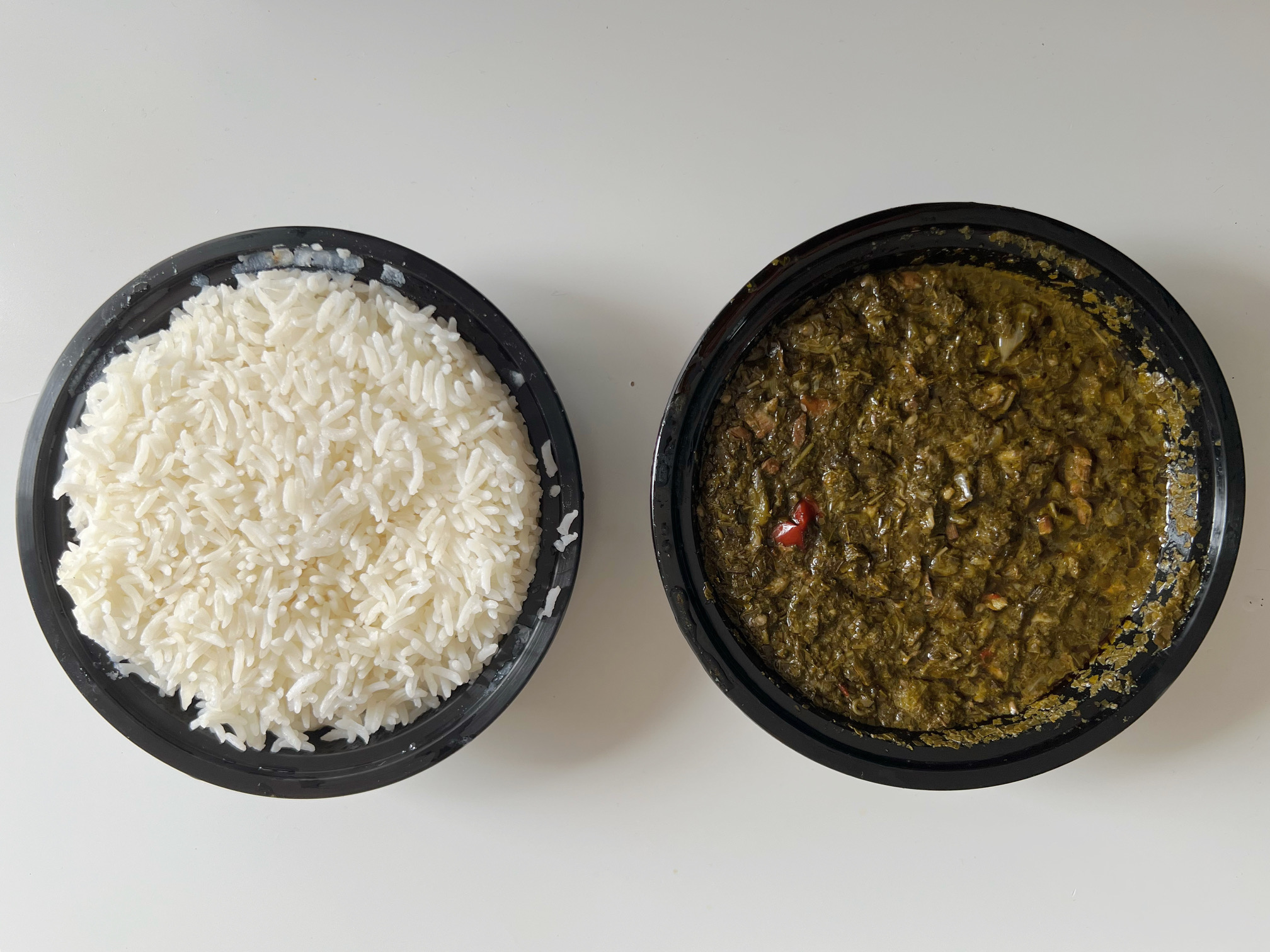 On a white table, there are two circular plastic containers. The one on the left is full of white rice, and the one one the right is full of pondu, stewed cassava leaves. Photo by Alyssa Buckley.