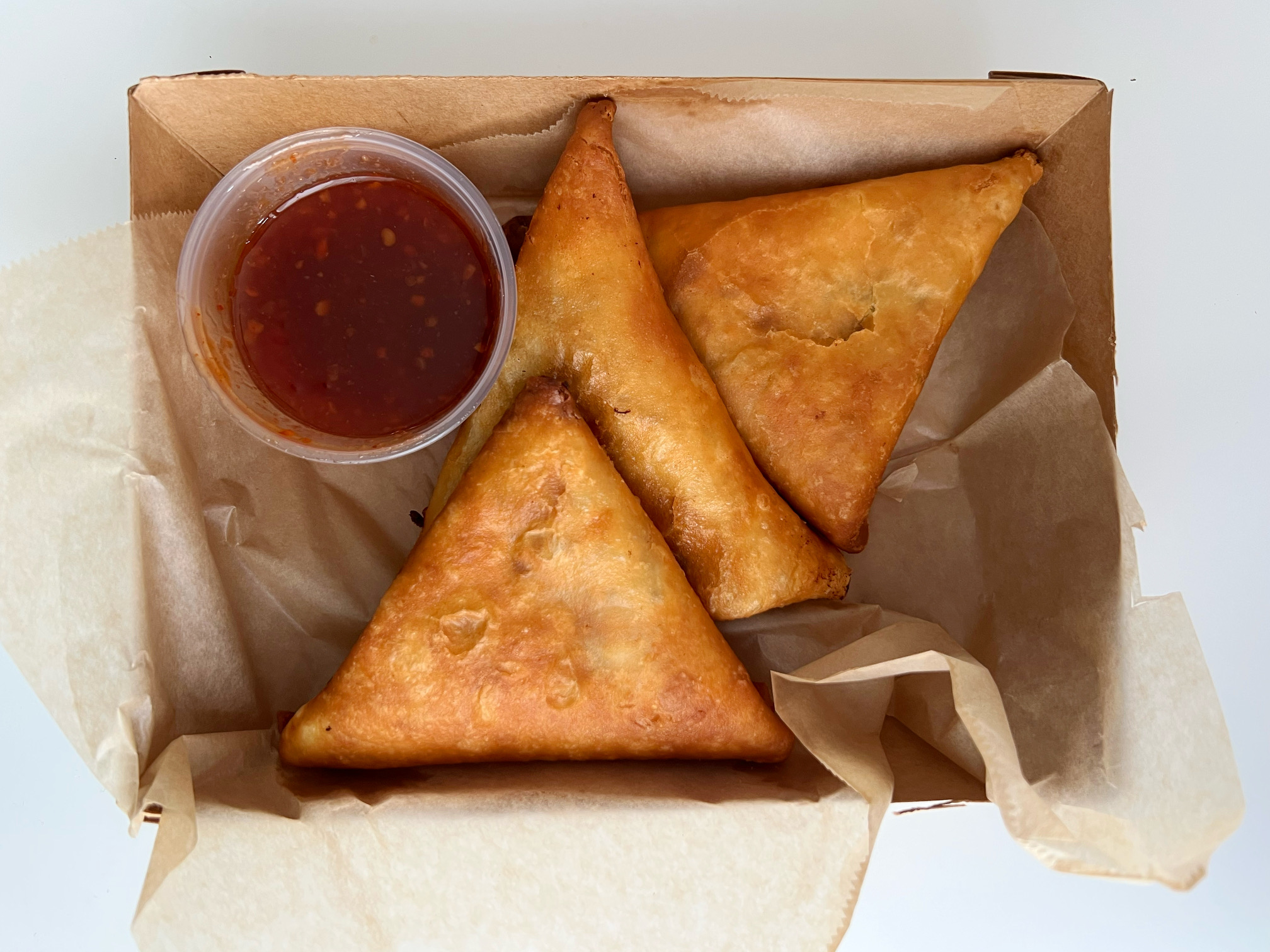 In a small cardboard box, there are three triangular samosas with a side cup of red chili sauce. Photo by Alyssa Buckley.