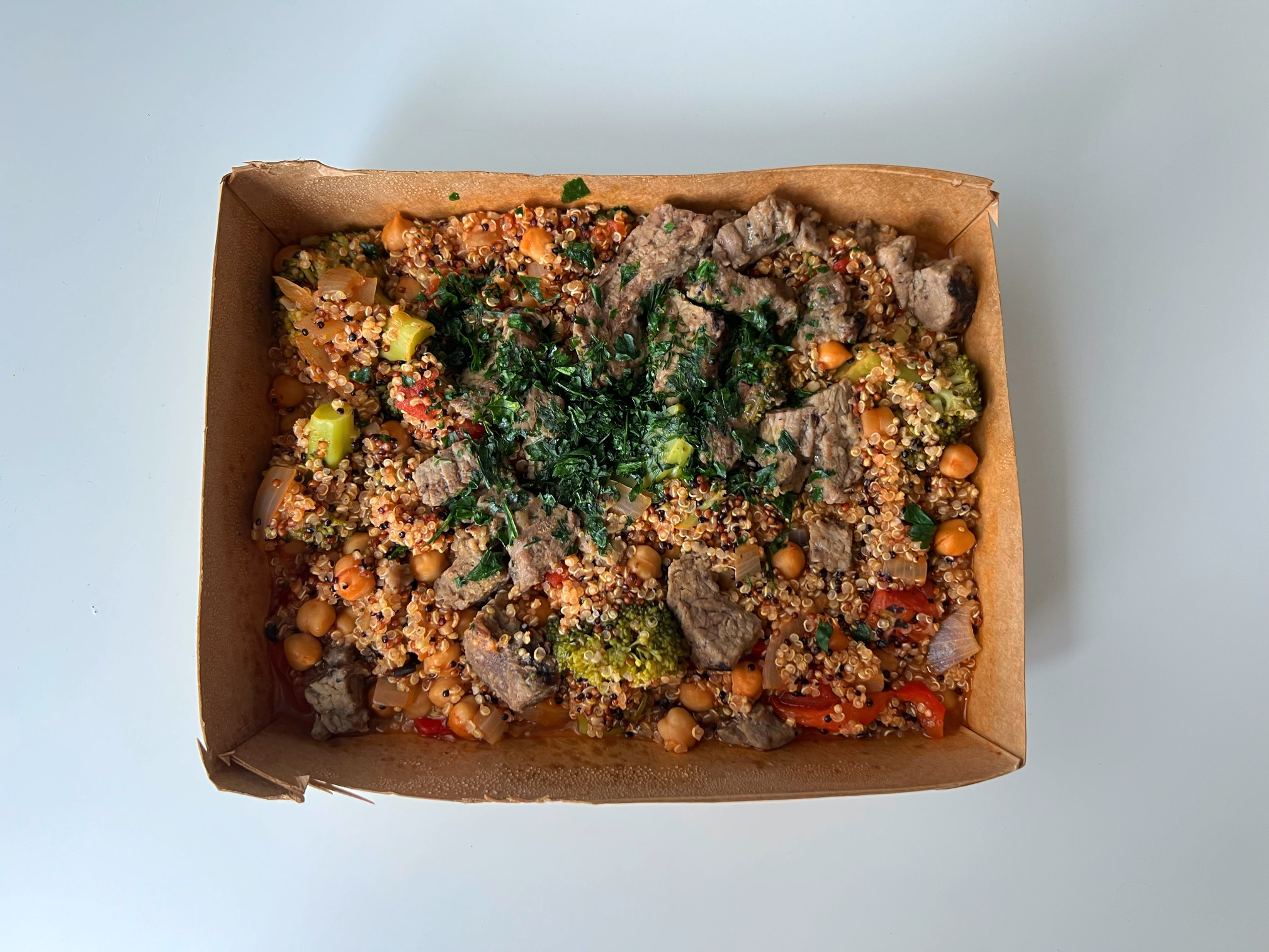 On a white table, there is a cardboard box with quinoa, beef, broccoli, chickpeas, and veggies topped with cilantro. Photo by Alyssa Buckley.