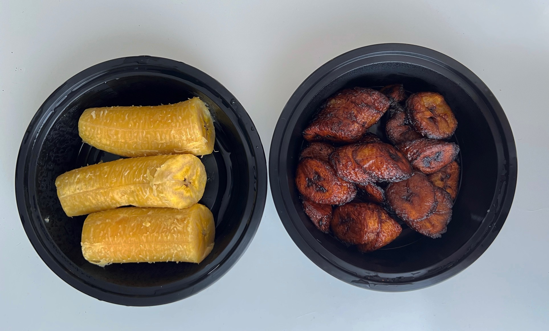 On a white table, there are two black circular containers with plantains. On the left, there are three peeled boiled plantains, and on the right, there are many dark brown medallions of fried plantains. Photo by Alyssa Buckley.