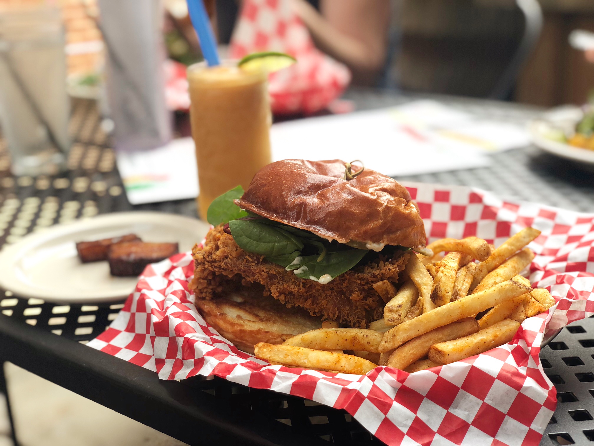 On a black patio table, there is a plate of food lined with a red-and-white checkered parchment paper. There is a fried chicken sandwich with fries and an orange cocktail behind it. Photo by Alyssa Buckley.