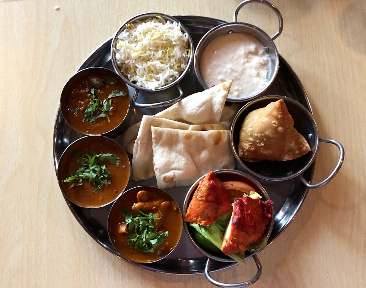 On a light wood table, there is a metal tray with small metal bowls of Indian dishes with four pieces of naan in the center. Photo by Alyssa Buckley.
