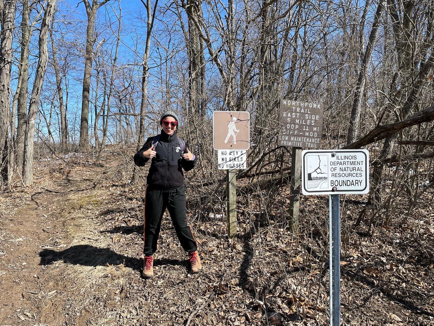 Photograph of author Mara Thacker standing next to three signs indicating the trailhead for the North Fork Trail. There are trees in the background, colors are mostly brown with a blue sky in the background.